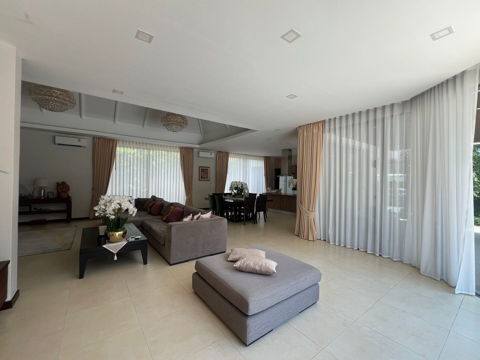 Spacious, modern living room with gray sectional sofa, glass coffee table, and white tiled floor; large windows with sheer curtains let in natural light.