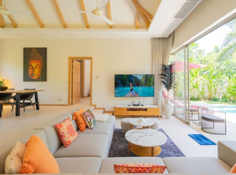 Bright living room with a beige sectional, orange cushions, and marble coffee tables facing a TV; glass doors overlook a pool and tropical garden.