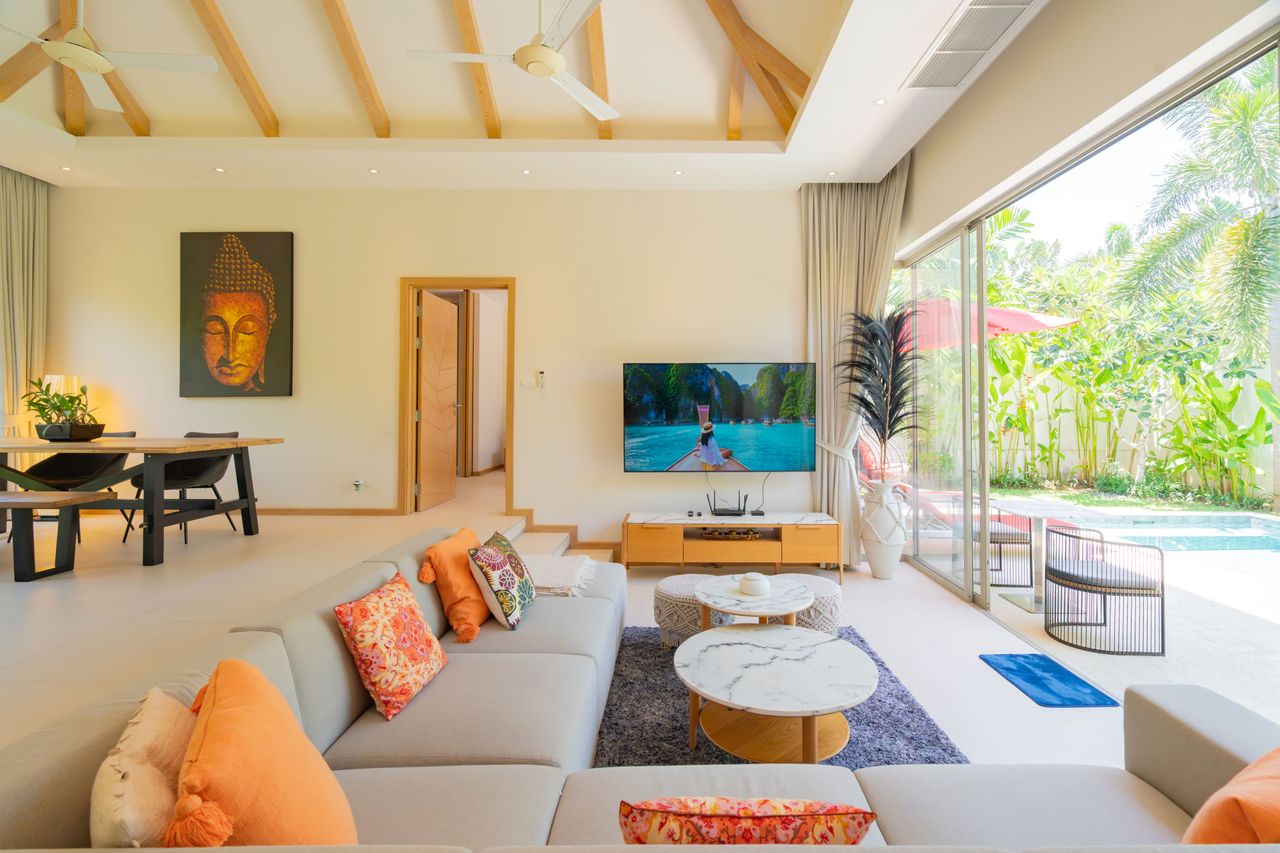 Bright living room with a beige sectional, orange cushions, and marble coffee tables facing a TV; glass doors overlook a pool and tropical garden.