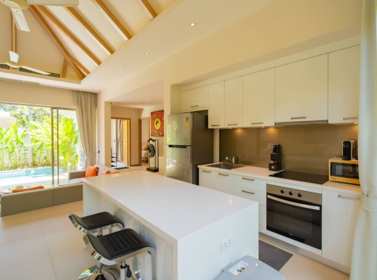 Open-plan kitchen with white island, bar stools, and stainless steel appliances adjacent to a living area and sliding glass doors to a lush outdoor pool area.