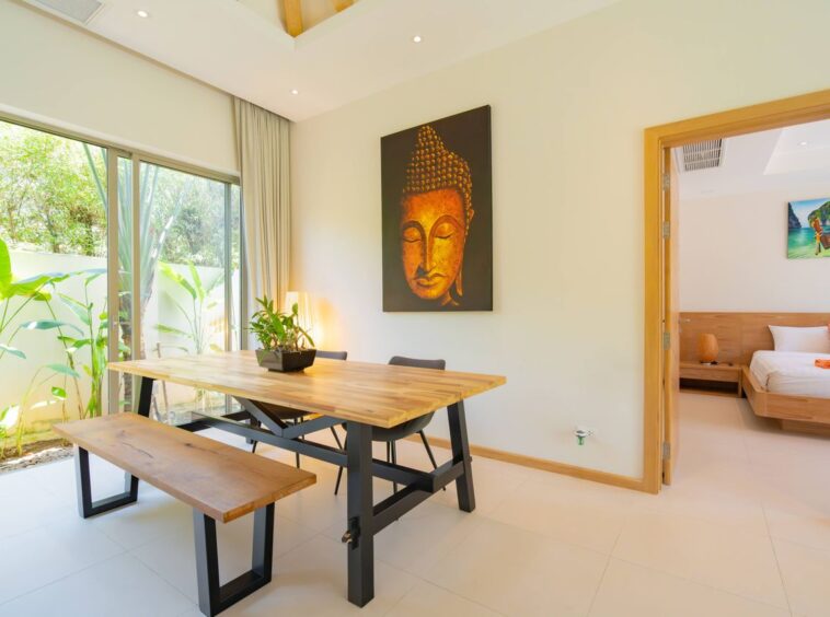 Bright dining area with a wooden table and bench, Buddha artwork on the wall, and sliding glass doors opening to greenery outside.