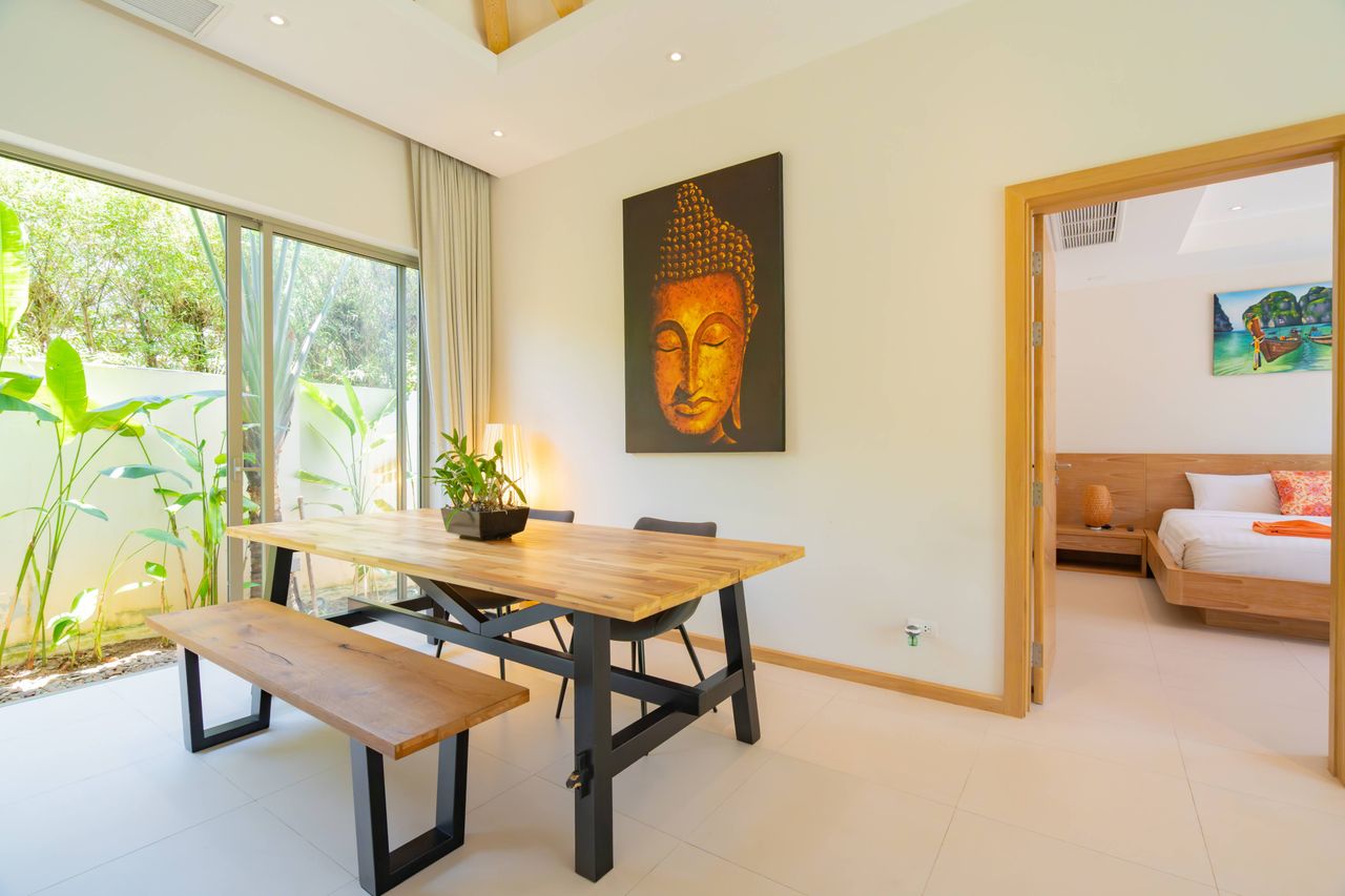 Bright dining area with a wooden table and bench, Buddha artwork on the wall, and sliding glass doors opening to greenery outside.