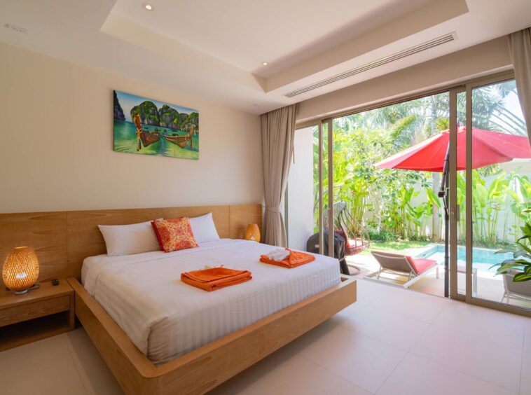 Bright hotel room with a wooden platform bed, white linens, orange towels, and orange pillow; sliding glass doors open to a tropical patio with a pool and red umbrella.
