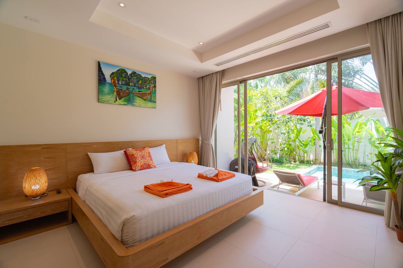 Bright hotel room with a wooden platform bed, white linens, orange towels, and orange pillow; sliding glass doors open to a tropical patio with a pool and red umbrella.