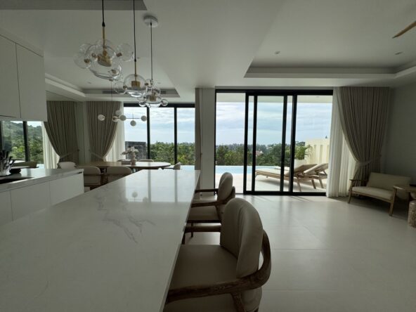 Open-plan kitchen and dining area with a white marble island, wicker chairs, and large glass doors opening to a pool and outdoor seating.