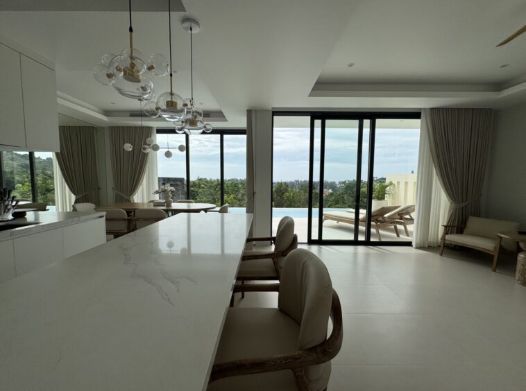 Open-plan kitchen and dining area with a white marble island, wicker chairs, and large glass doors opening to a pool and outdoor seating.