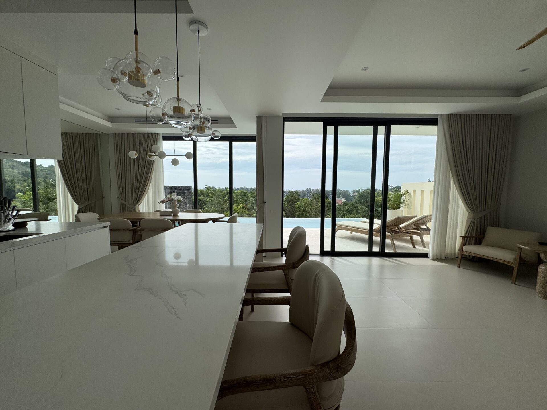 Open-plan kitchen and dining area with a white marble island, wicker chairs, and large glass doors opening to a pool and outdoor seating.