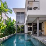 Modern two-story villa with a rectangular pool, lush tropical plants along the left, and an outdoor dining area under a covered patio.