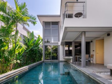 Modern two-story villa with a rectangular pool, lush tropical plants along the left, and an outdoor dining area under a covered patio.
