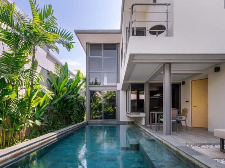 Modern two-story villa with a rectangular pool, lush tropical plants along the left, and an outdoor dining area under a covered patio.