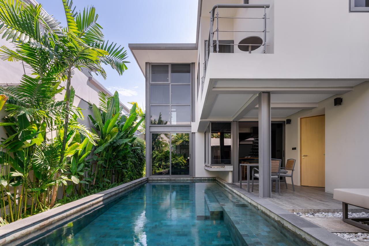 Modern two-story villa with a rectangular pool, lush tropical plants along the left, and an outdoor dining area under a covered patio.