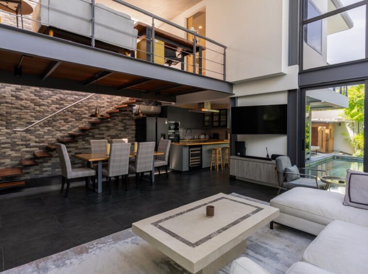 Open-plan living area with dining table, kitchen, and a wooden staircase beneath a mezzanine balcony.