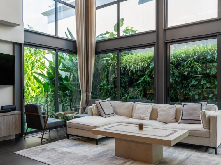 Bright contemporary living room with floor-to-ceiling glass walls, beige sectional sofa, and lush tropical plants outside.