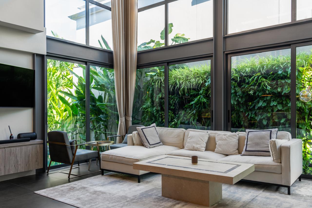 Bright contemporary living room with floor-to-ceiling glass walls, beige sectional sofa, and lush tropical plants outside.