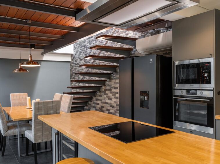 Open-concept kitchen with a wooden island, black induction cooktop, and stainless steel fridge and ovens beside a floating wooden staircase against a brick wall.