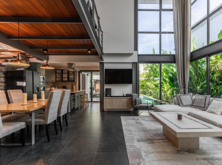 Open-plan living and dining area with a long wood dining table, upholstered chairs, a modern kitchen, and seating by large windows.