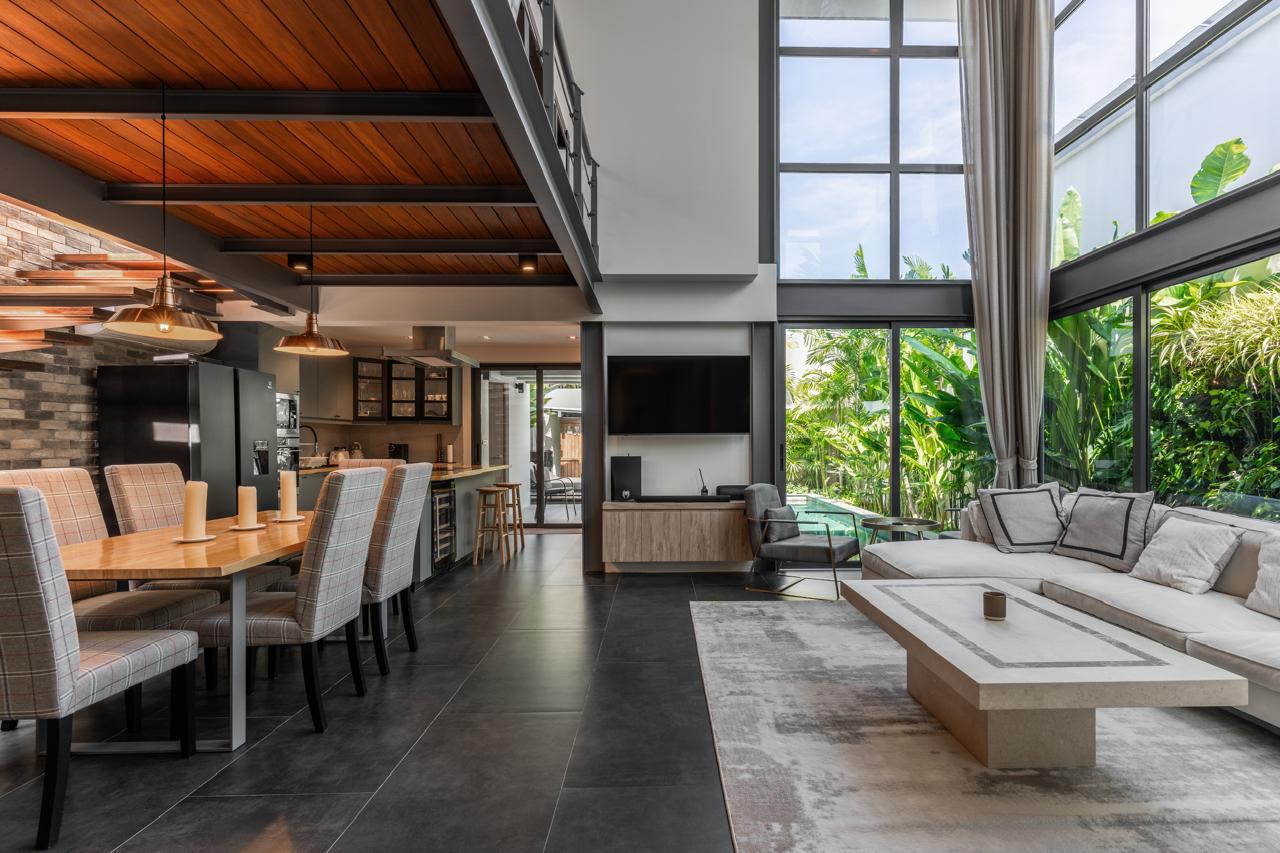 Open-plan living and dining area with a long wood dining table, upholstered chairs, a modern kitchen, and seating by large windows.
