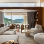 Luxurious living room with cream sectional sofas, curved chairs, and wood-paneled walls framing a glass door to an outdoor balcony overlooking a coastal town and green hills.