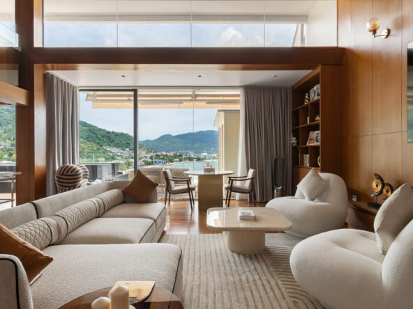 Luxurious living room with cream sectional sofas, curved chairs, and wood-paneled walls framing a glass door to an outdoor balcony overlooking a coastal town and green hills.