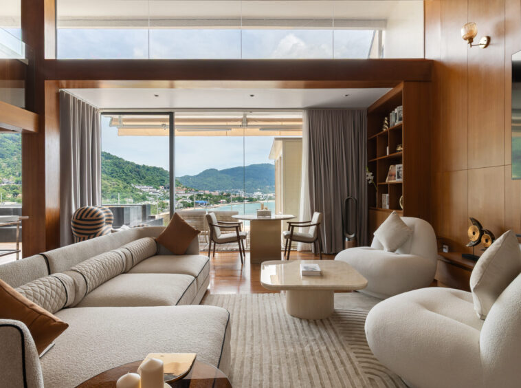 Luxurious living room with cream sectional sofas, curved chairs, and wood-paneled walls framing a glass door to an outdoor balcony overlooking a coastal town and green hills.