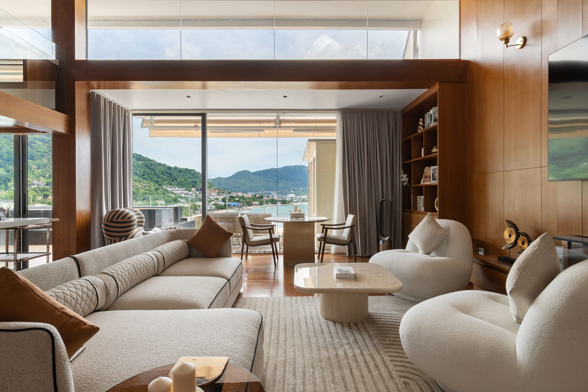 Luxurious living room with cream sectional sofas, curved chairs, and wood-paneled walls framing a glass door to an outdoor balcony overlooking a coastal town and green hills.