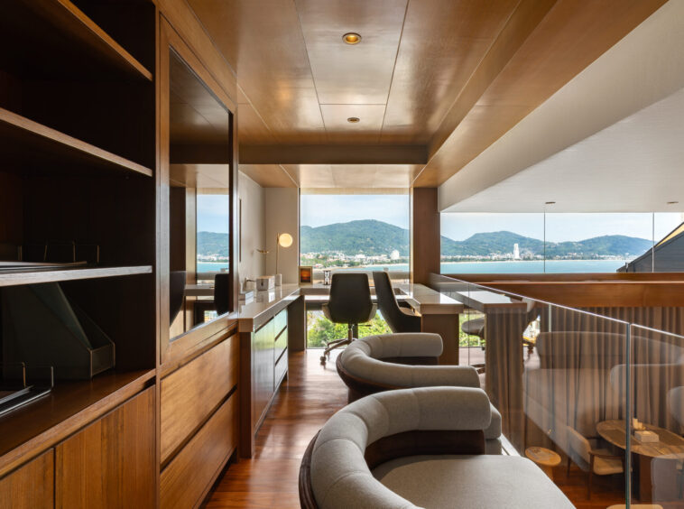 Luxurious wooden office with curved gray chairs and a glass railing, overlooking a bay and mountains through large windows.