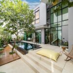 Modern two-story home courtyard with a narrow pool, glass walls, and lounge seating amid tropical plants.