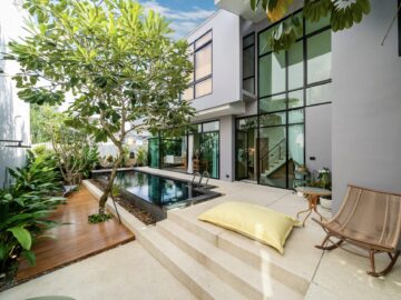 Modern two-story home courtyard with a narrow pool, glass walls, and lounge seating amid tropical plants.