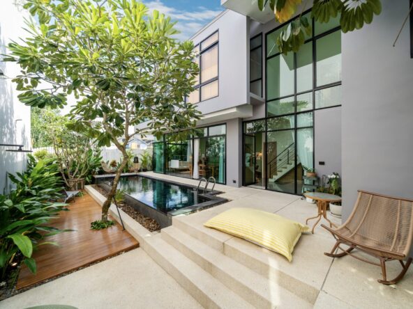 Modern two-story home courtyard with a narrow pool, glass walls, and lounge seating amid tropical plants.