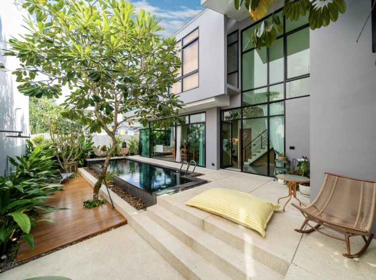 Modern two-story home courtyard with a narrow pool, glass walls, and lounge seating amid tropical plants.