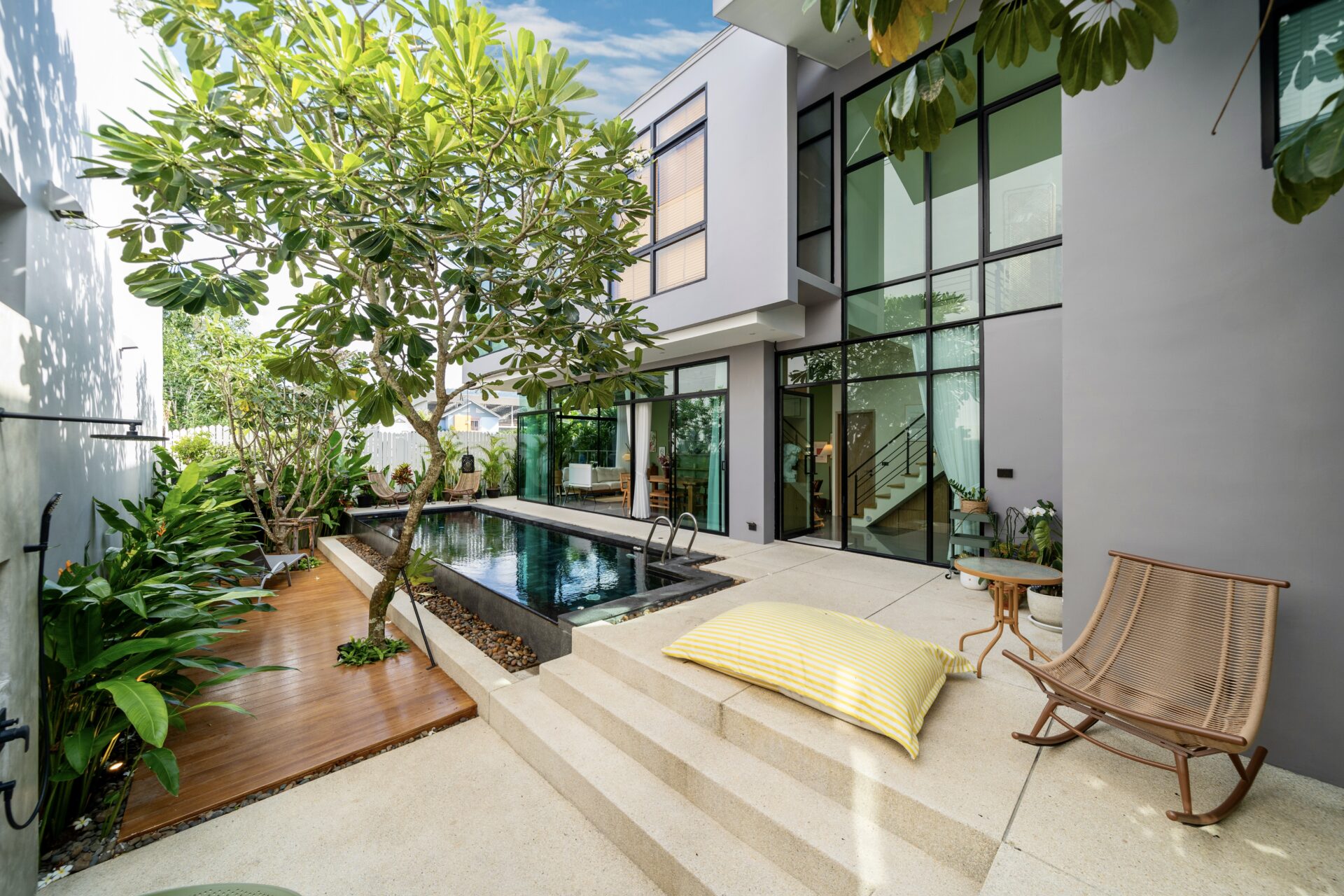 Modern two-story home courtyard with a narrow pool, glass walls, and lounge seating amid tropical plants.