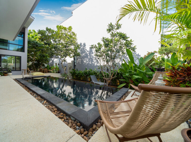 Modern courtyard with a rectangular pool, tan deck, and wicker lounge chairs amid tropical plants by a white building.