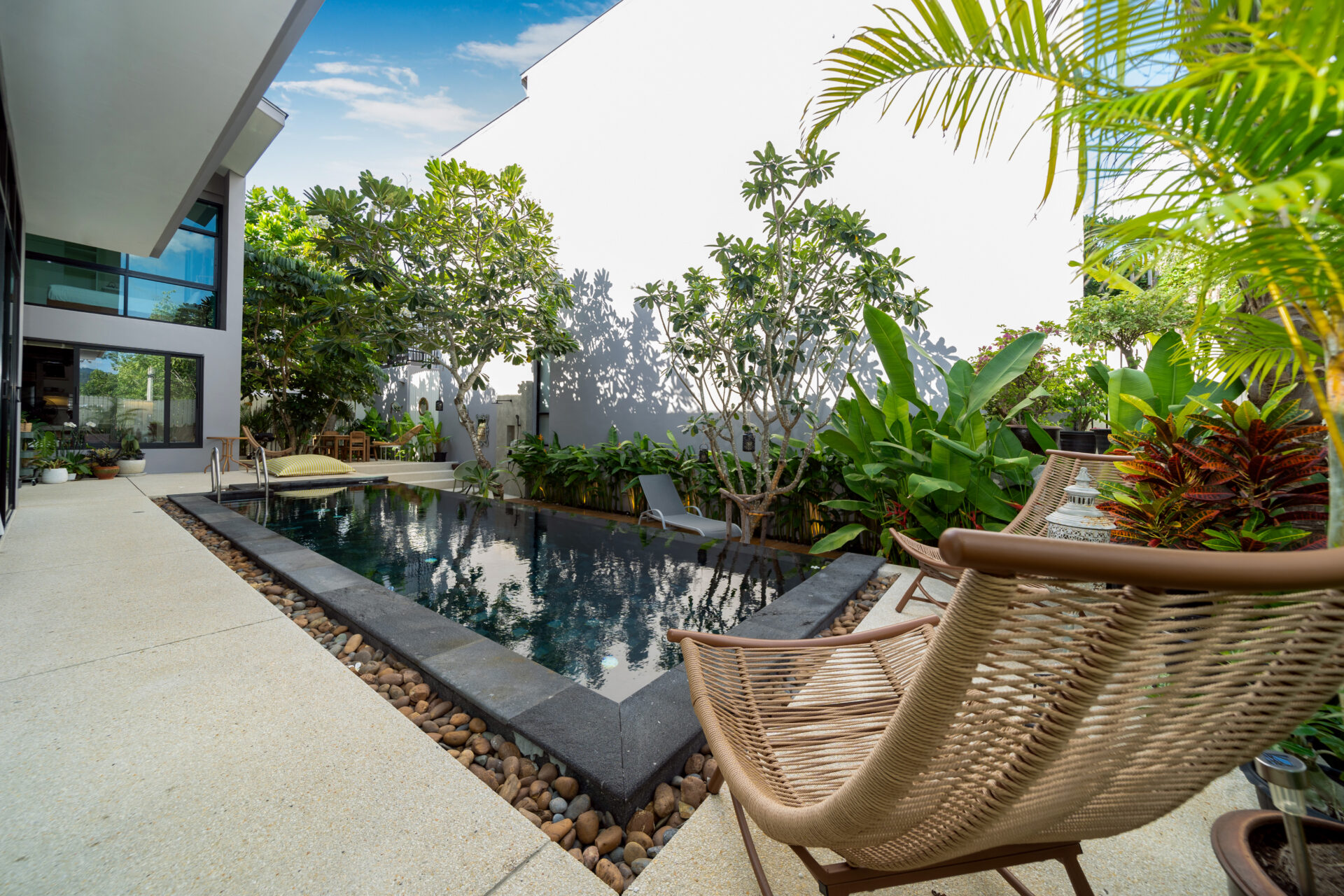Modern courtyard with a rectangular pool, tan deck, and wicker lounge chairs amid tropical plants by a white building.