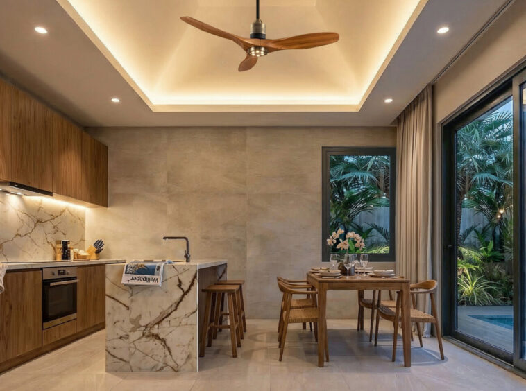 Modern open-plan kitchen and dining area with wood cabinets, marble island, and warm recessed ceiling lighting overlooking a tropical garden and pool through large glass doors