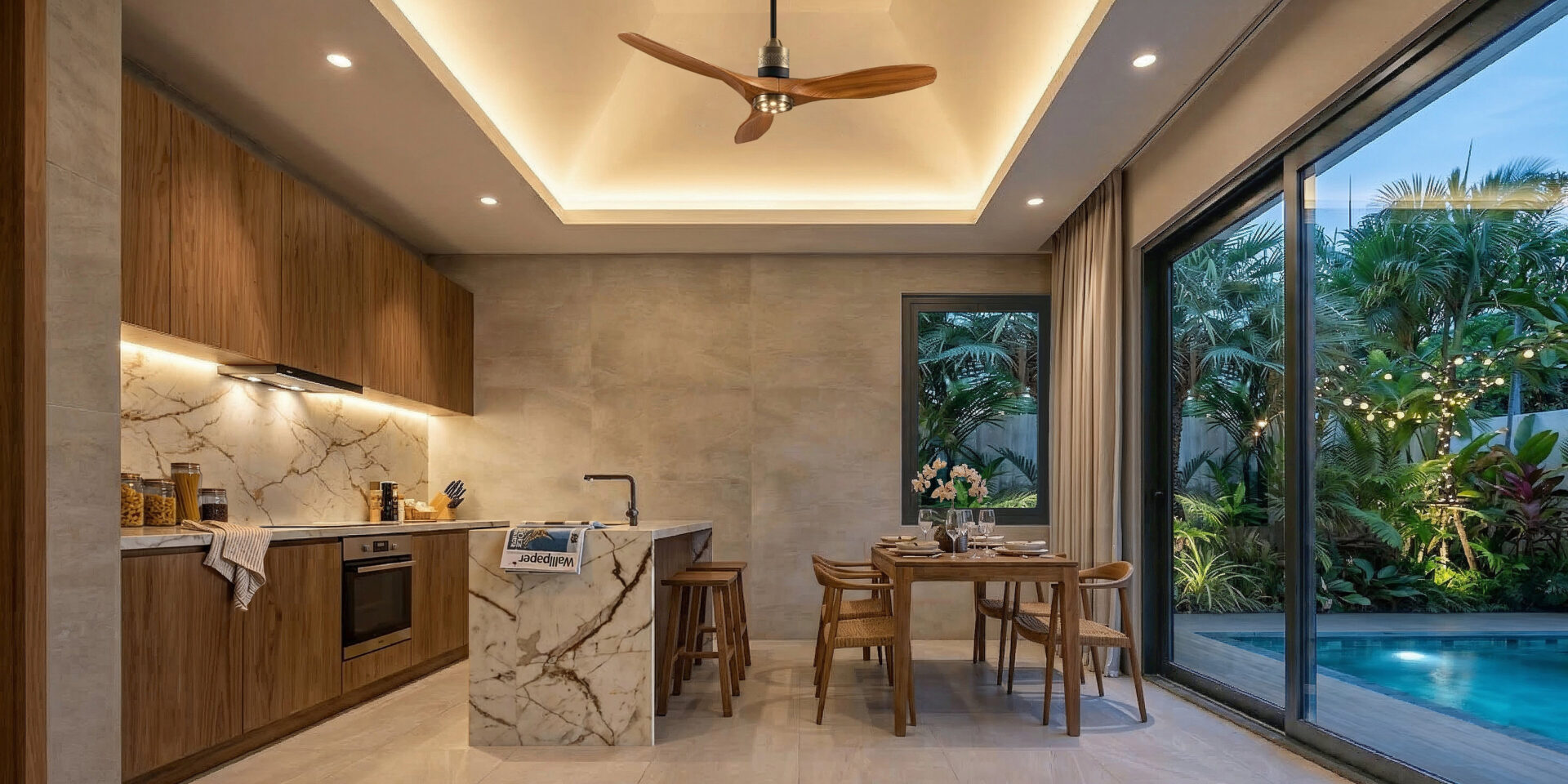 Modern open-plan kitchen and dining area with wood cabinets, marble island, and warm recessed ceiling lighting overlooking a tropical garden and pool through large glass doors