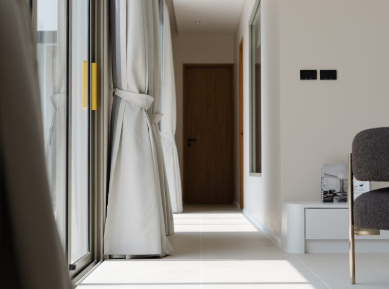 Bright modern hallway with floor-to-ceiling windows and tied-back curtains on the left, leading to a wooden door at the end.