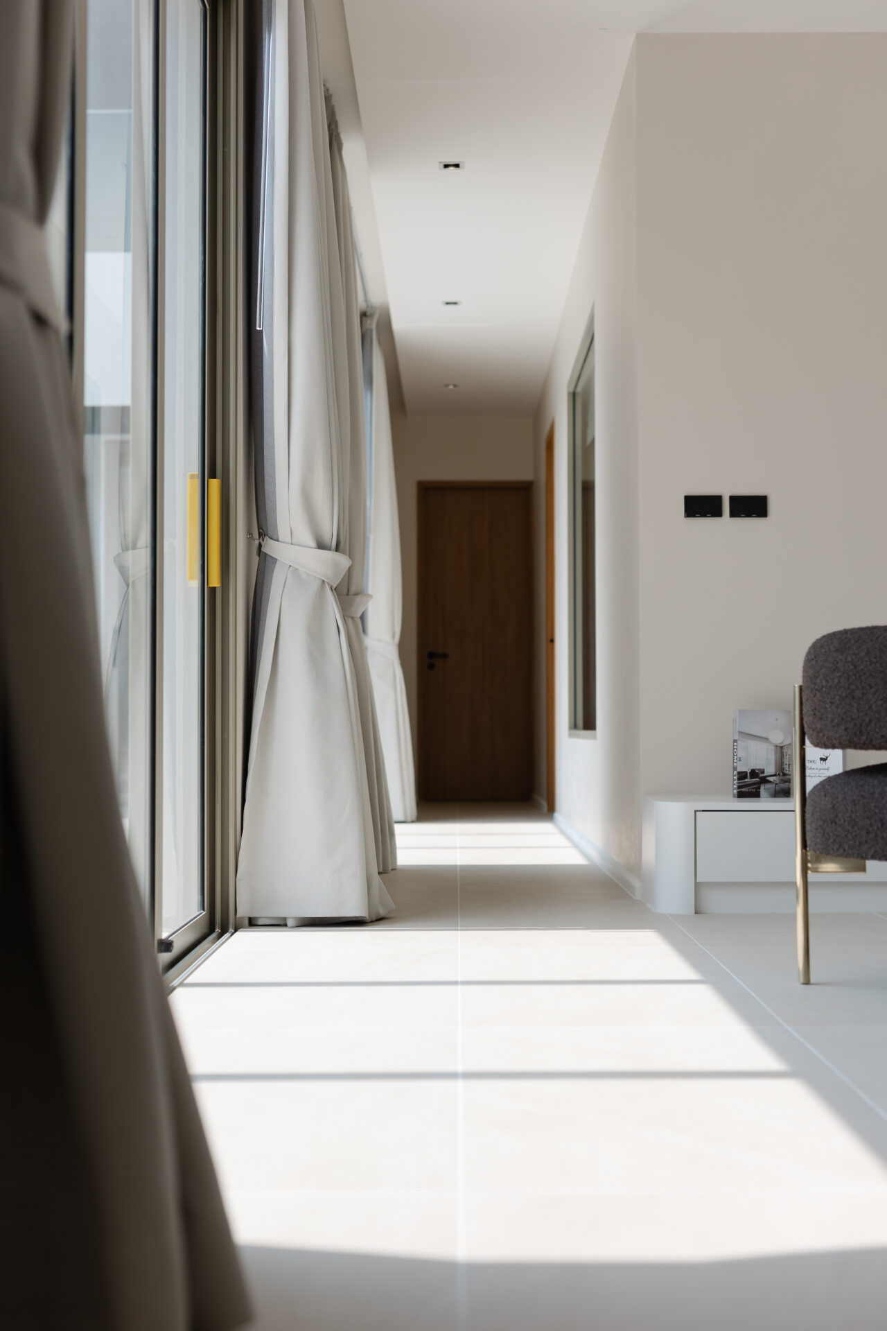 Bright modern hallway with floor-to-ceiling windows and tied-back curtains on the left, leading to a wooden door at the end.