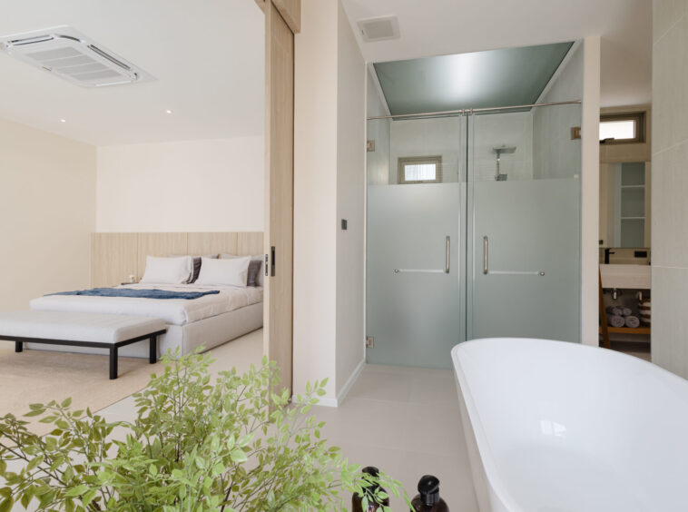 Open-plan hotel suite showing a bed area adjacent to a glass-walled bathroom with a frosted lower panel and a freestanding bathtub in the foreground, plus green plants in front.