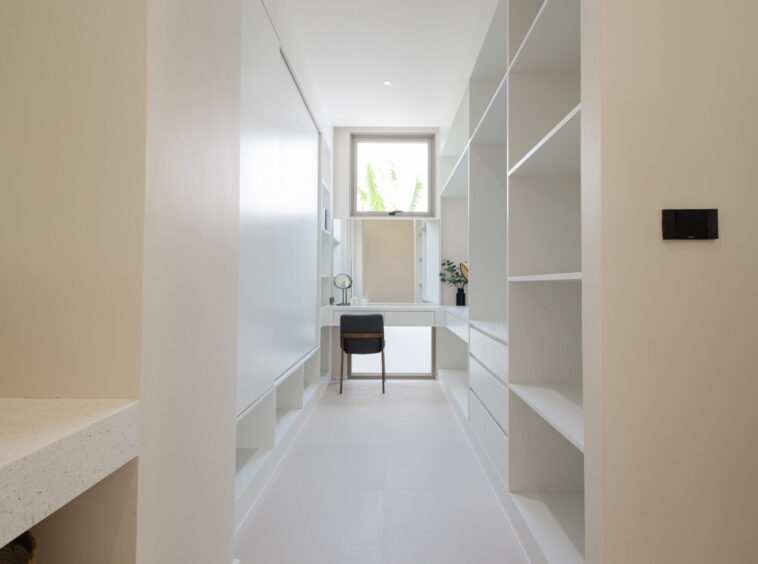 Bright walk-in closet with white built-in shelves, a desk, and a window at the far end letting in light.