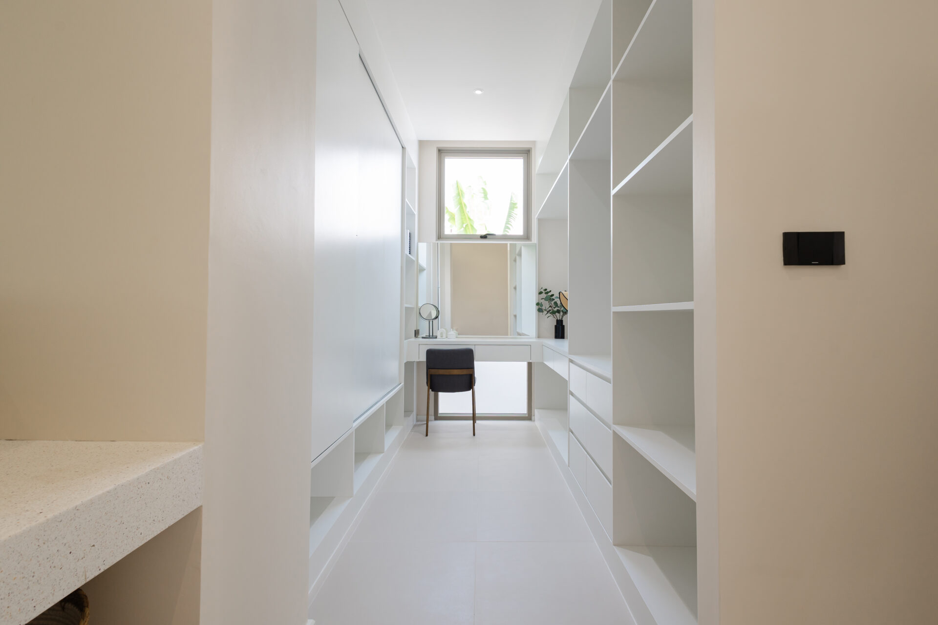 Bright walk-in closet with white built-in shelves, a desk, and a window at the far end letting in light.