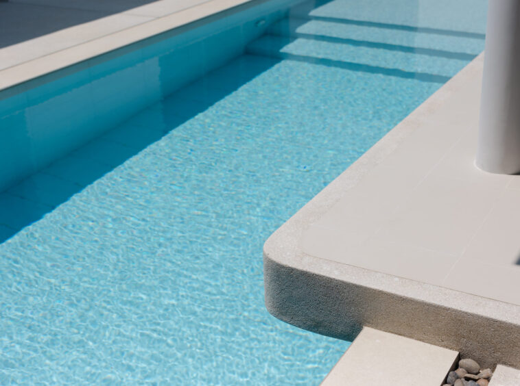 Pool edge beside a clear blue pool with stepped entry and a light gray tiled deck.