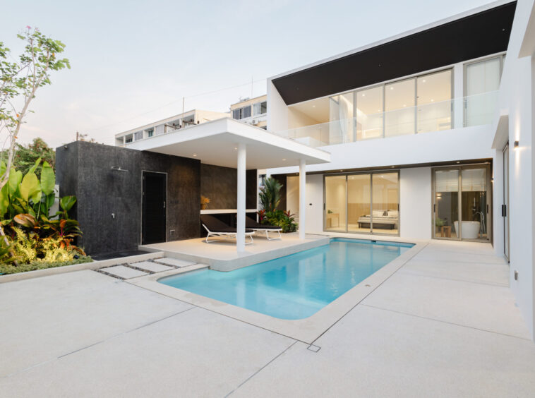Contemporary two-story home with glass walls, a rectangular pool, and white patio furniture. Lush plants line the left side.