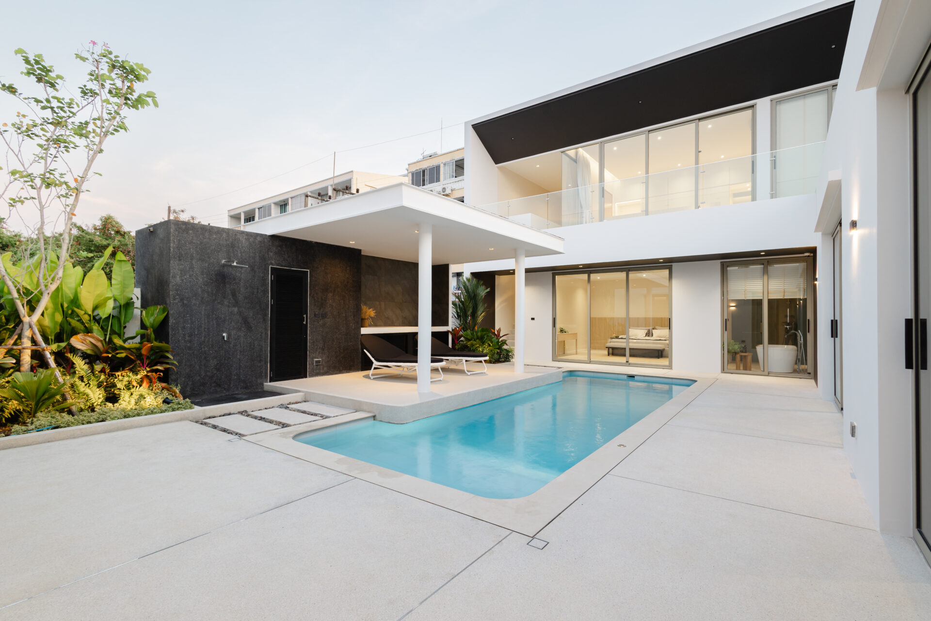Contemporary two-story home with glass walls, a rectangular pool, and white patio furniture. Lush plants line the left side.