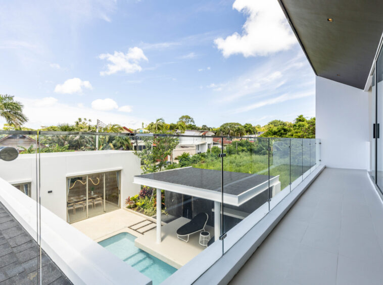 Contemporary balcony with glass railing and gray tile, overlooking a pool and tropical landscaping beneath a sunny sky.
