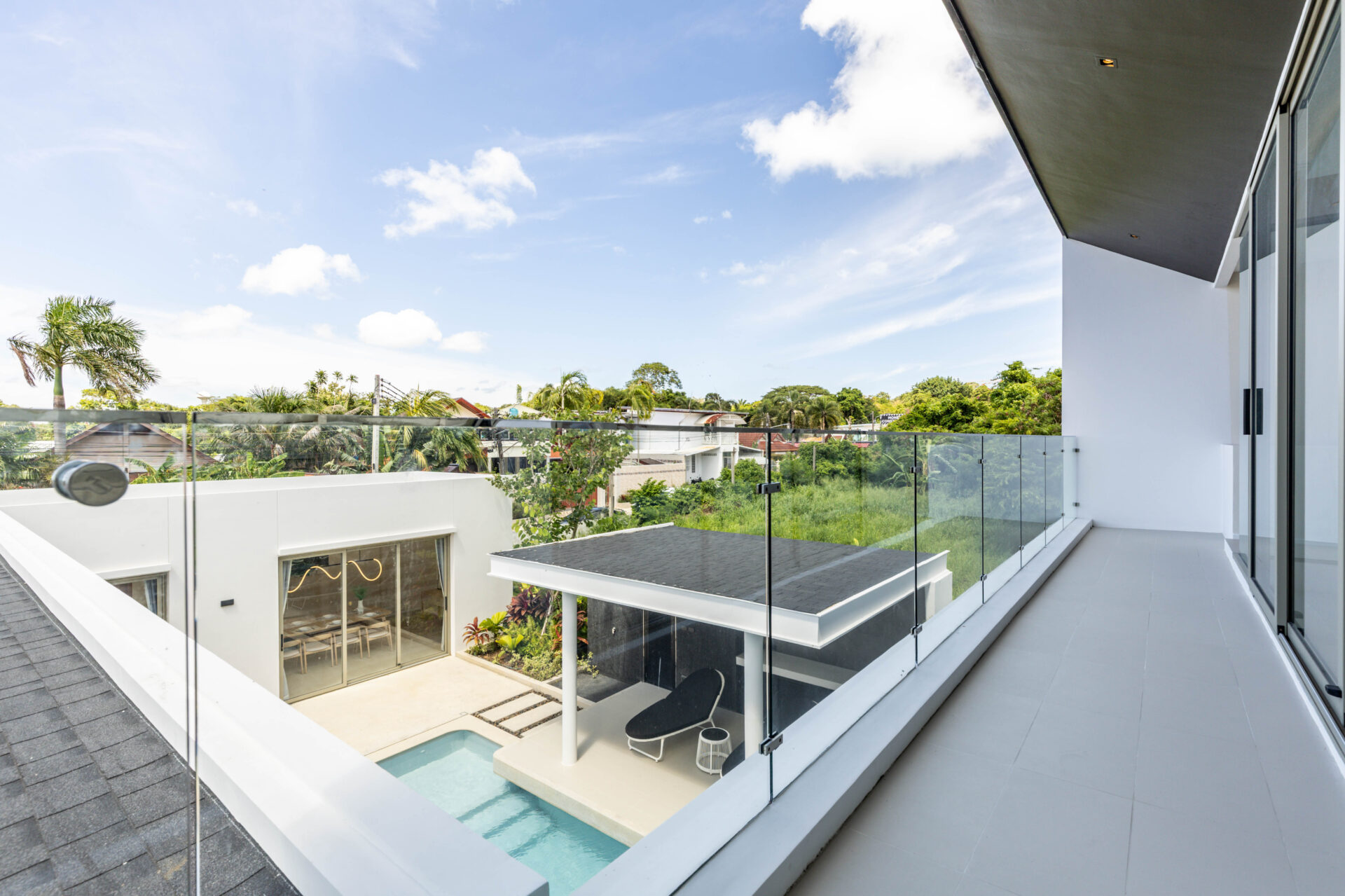 Contemporary balcony with glass railing and gray tile, overlooking a pool and tropical landscaping beneath a sunny sky.