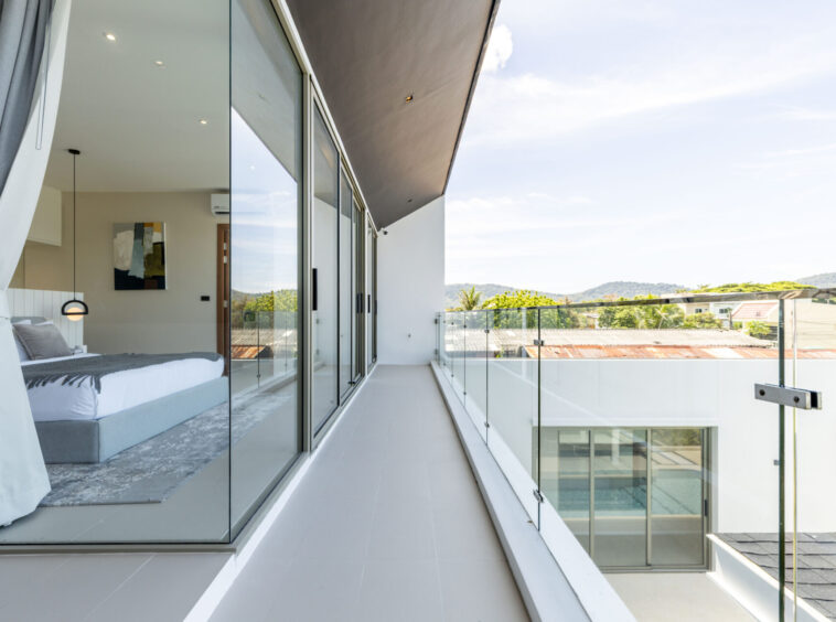 Modern balcony with glass railing and sliding glass doors; bed and bedside pendant visible inside, and a hills view beyond the balcony.