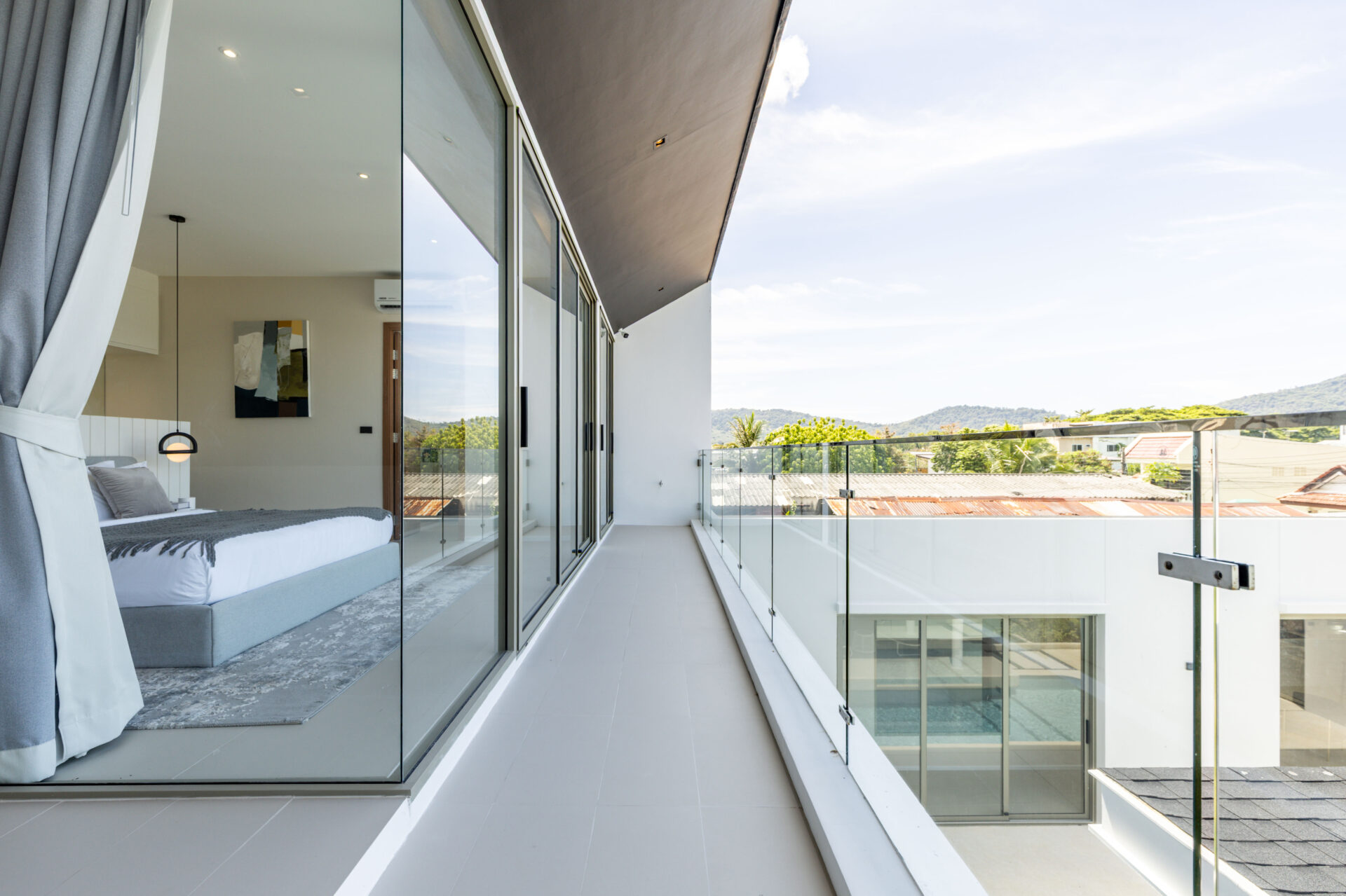Modern balcony with glass railing and sliding glass doors; bed and bedside pendant visible inside, and a hills view beyond the balcony.