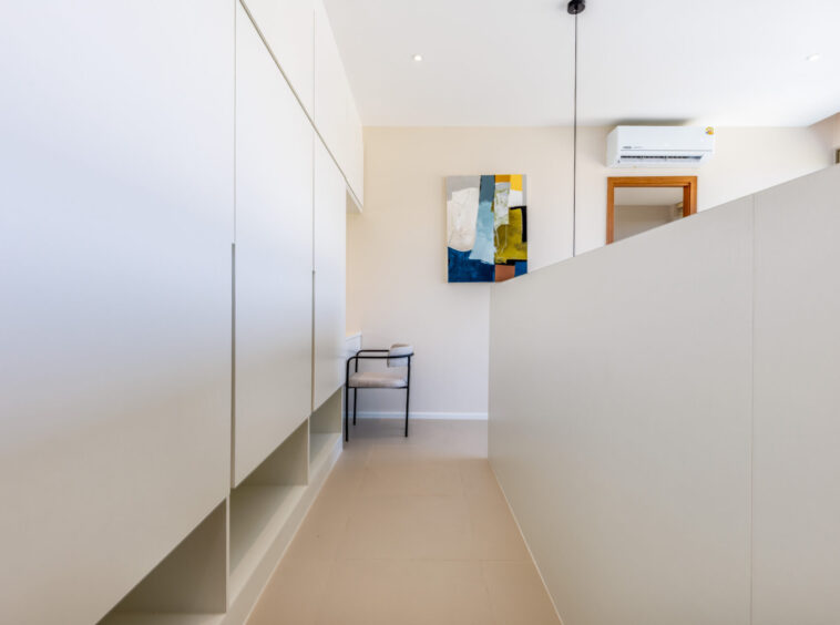 Narrow, bright hallway with white built-in cabinets on the left, beige walls, a colorful abstract painting, and a small metal chair beneath it.