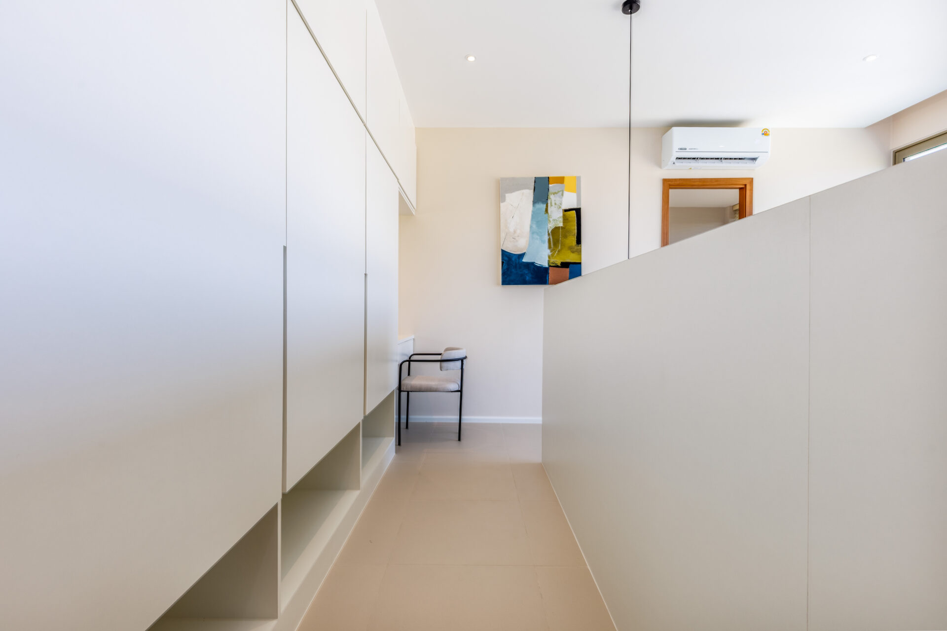 Narrow, bright hallway with white built-in cabinets on the left, beige walls, a colorful abstract painting, and a small metal chair beneath it.