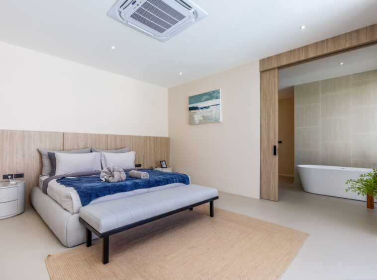 Modern hotel bedroom with a king bed, beige headboard, blue throw, and an open doorway to a tiled bathroom with a freestanding tub.